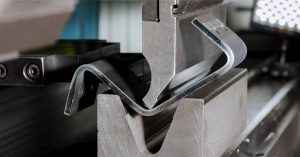 Close-up of press brake tool bending a thick piece of sheet metal by forcing it into a V-shaped die, creating a piece with several bends and angles.