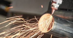 A close-up of a laser cutting machine cutting through a tube with orange streams of sparks flying out of the tube opening.